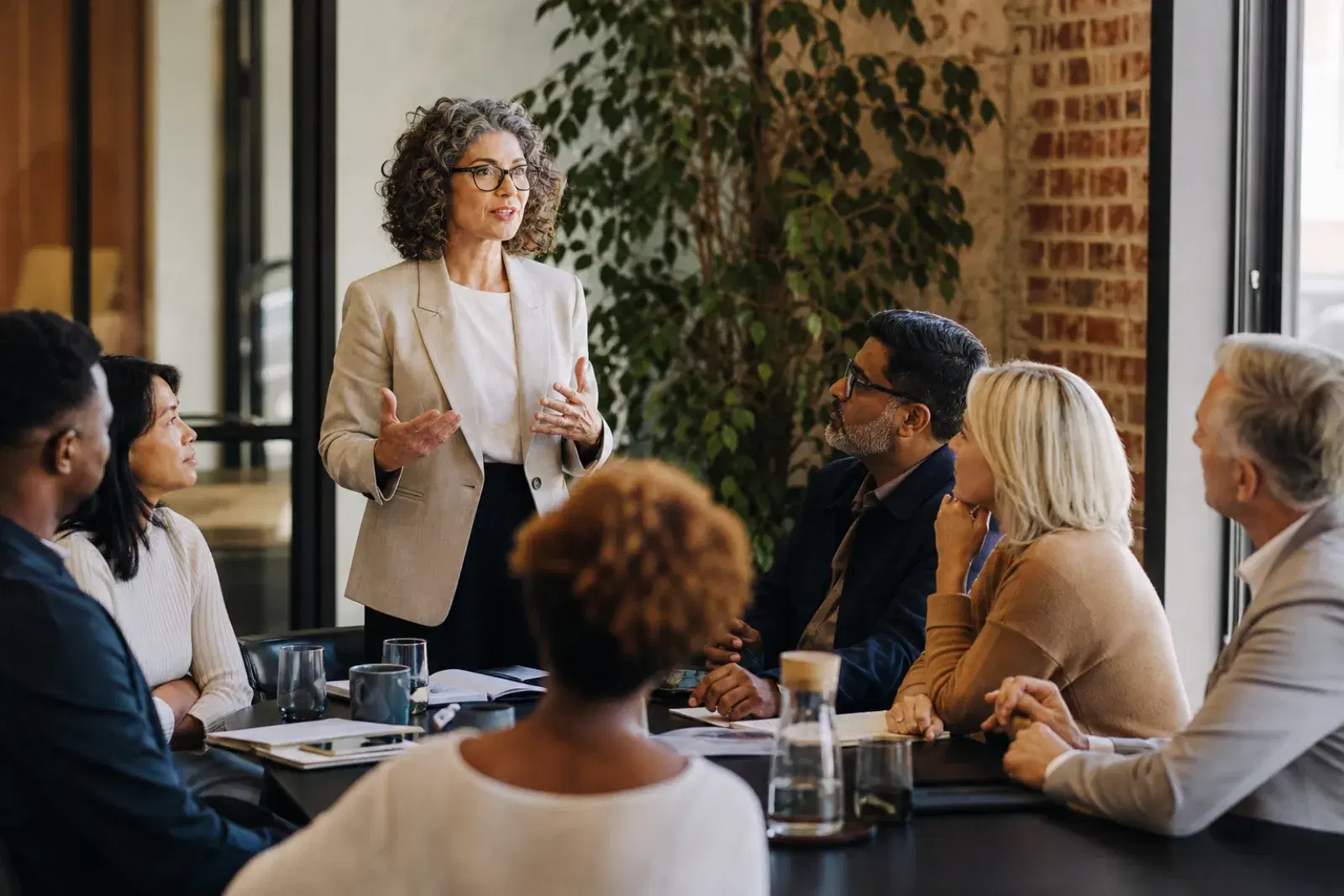 A leader standing at the head of a boardroom table with six colleagues turned toward her, listening.